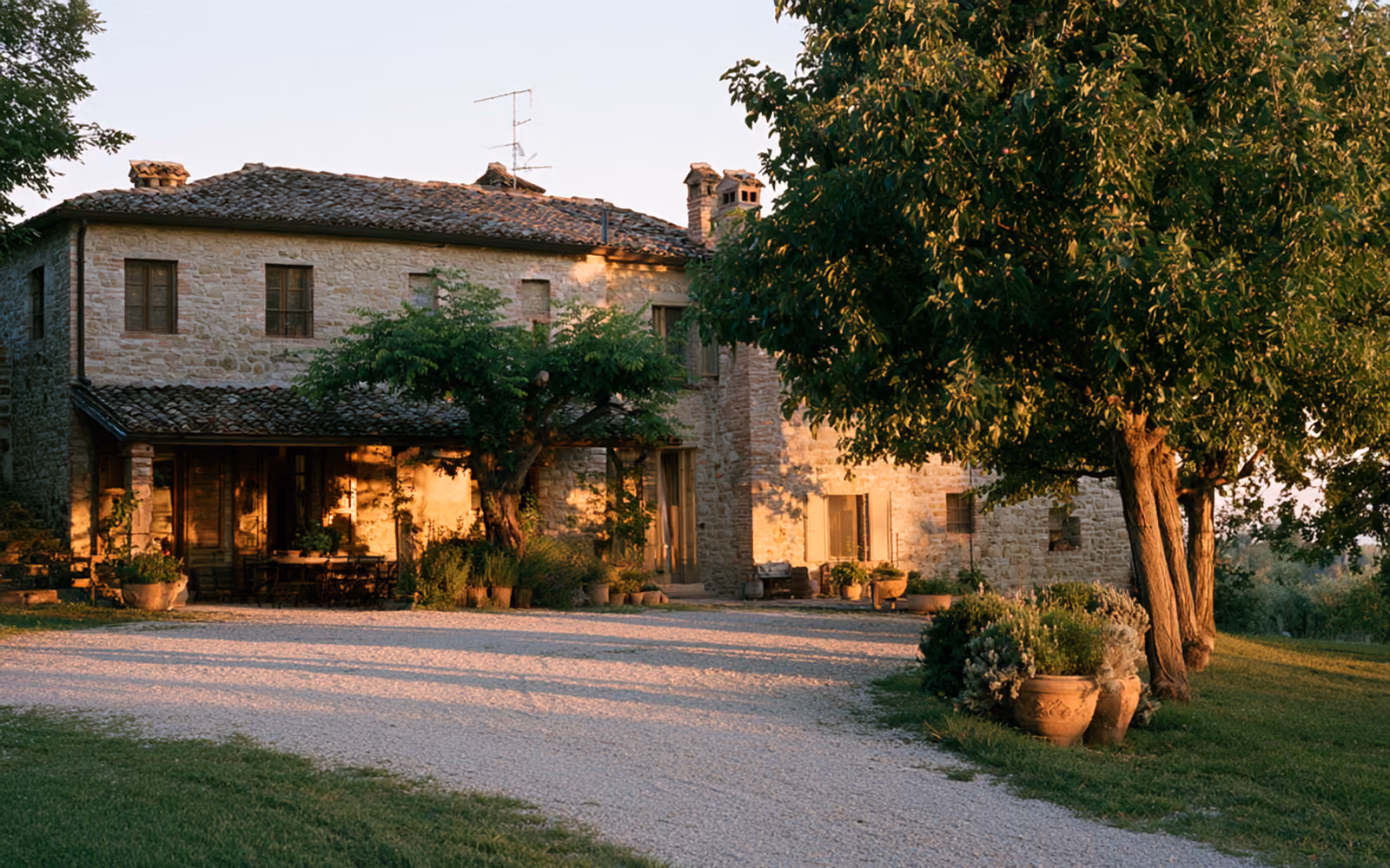 Casale in pietra emiliano al tramonto, con cortile in ghiaia e alberi accanto all'ingresso